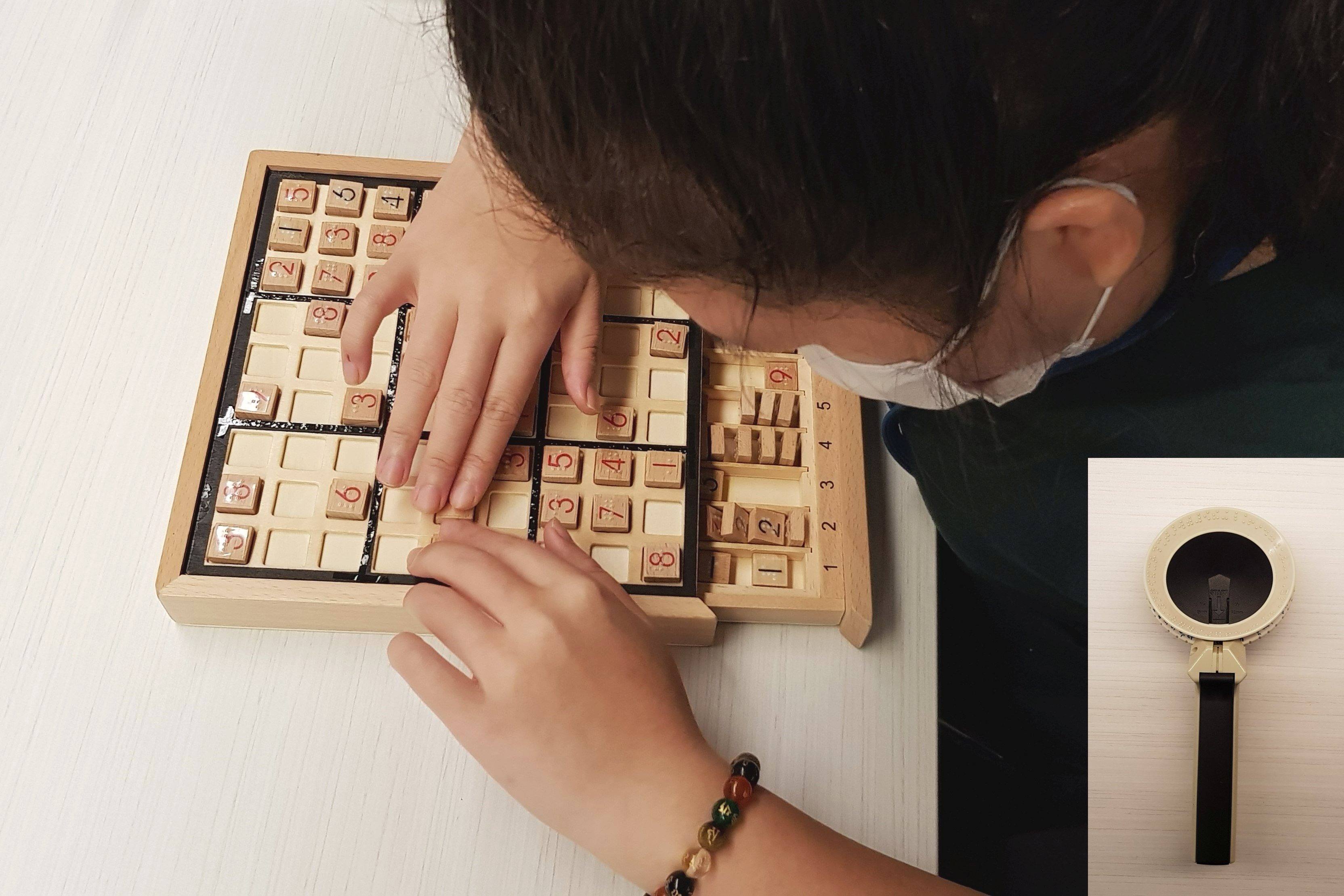 Photo of Siew Ling attempting to solve the Sudoku with the Wooden Sudoku set with drawer, and feeling the braille labelled on one of the number tiles.