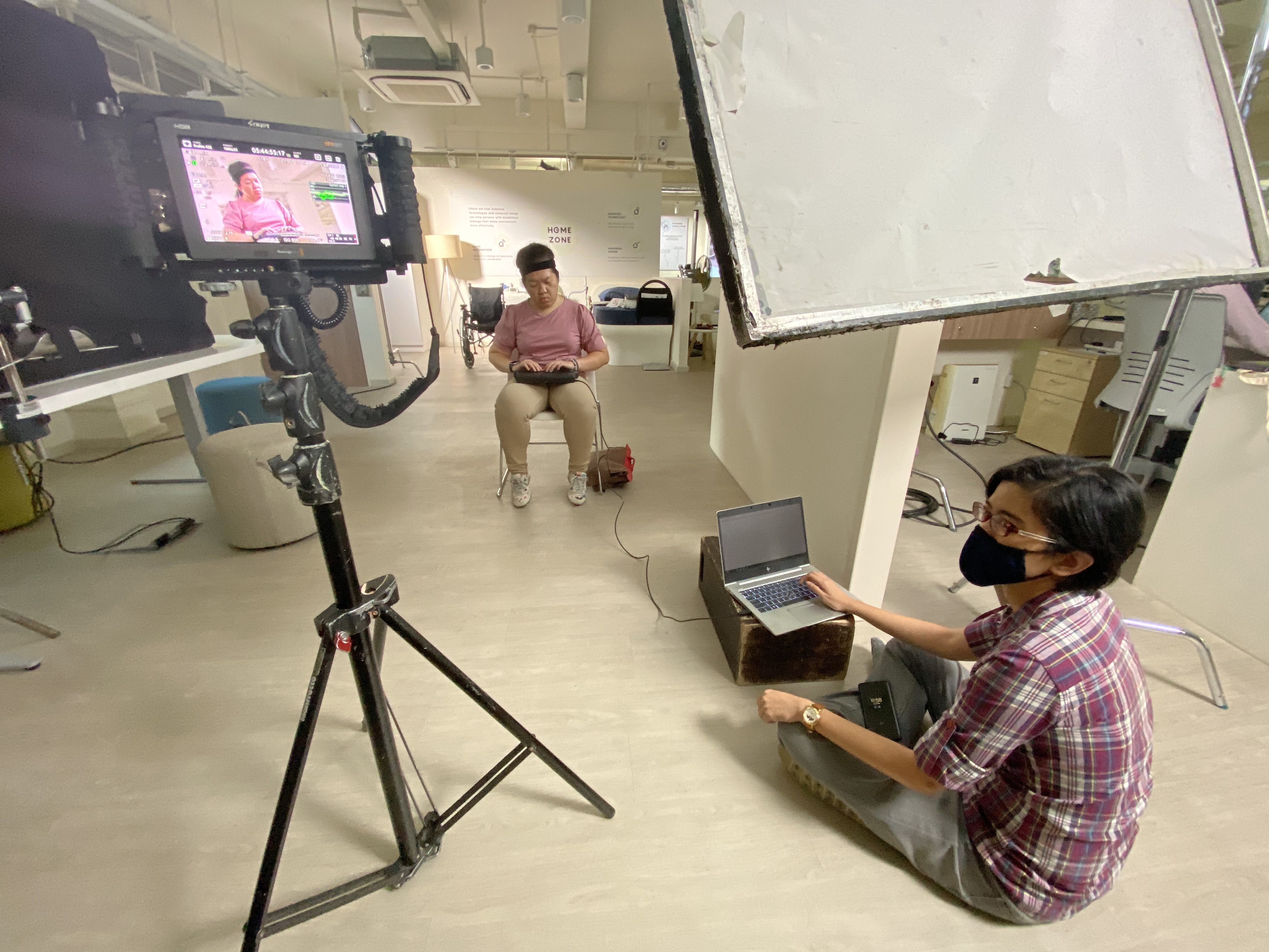 Siew Ling and colleague on filming set. Siew Ling is patiently reading her braille display while her colleague is observing production crew’s exchange off-frame.